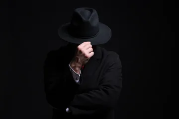 Pessoa de terno escuro e chapéu preto cobrindo o rosto, em fundo escuro, representando o black hat SEO