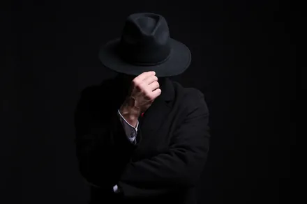 Pessoa de terno escuro e chapéu preto cobrindo o rosto, em fundo escuro, representando o black hat SEO