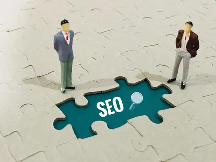 Dois bonecos de empresários sobre um quebra-cabeça com uma peça faltando que revela a palavra SEO e uma lupa